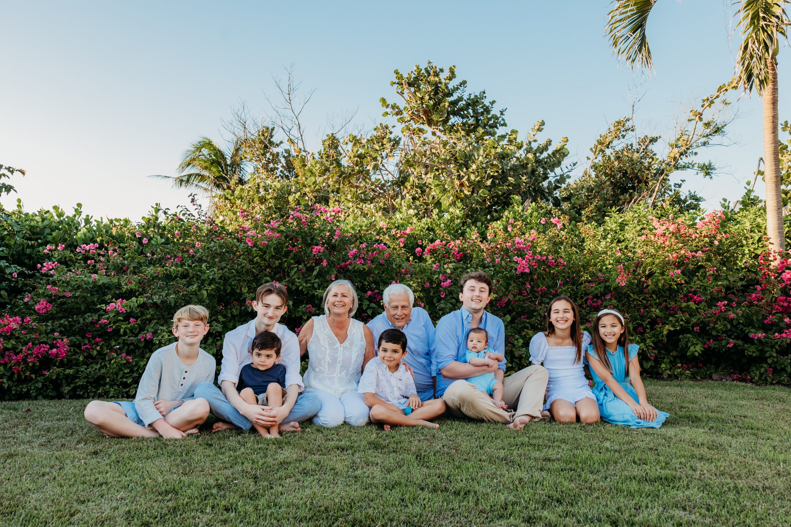 Grandparents seated on the grass with all nine of their grandchildren in front of a blooming bougainvillea hedge in Naples, Florida