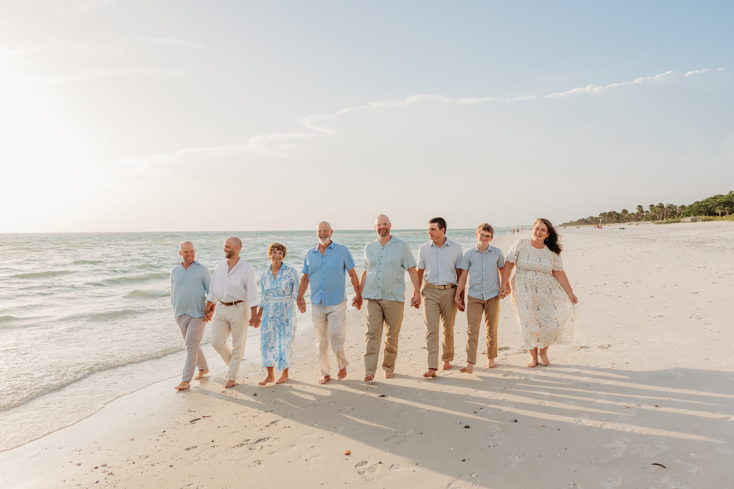 An extended family walking hand in hand along the shoreline at sunset in Naples, Florida, three generations dressed in coordinated blues and creams