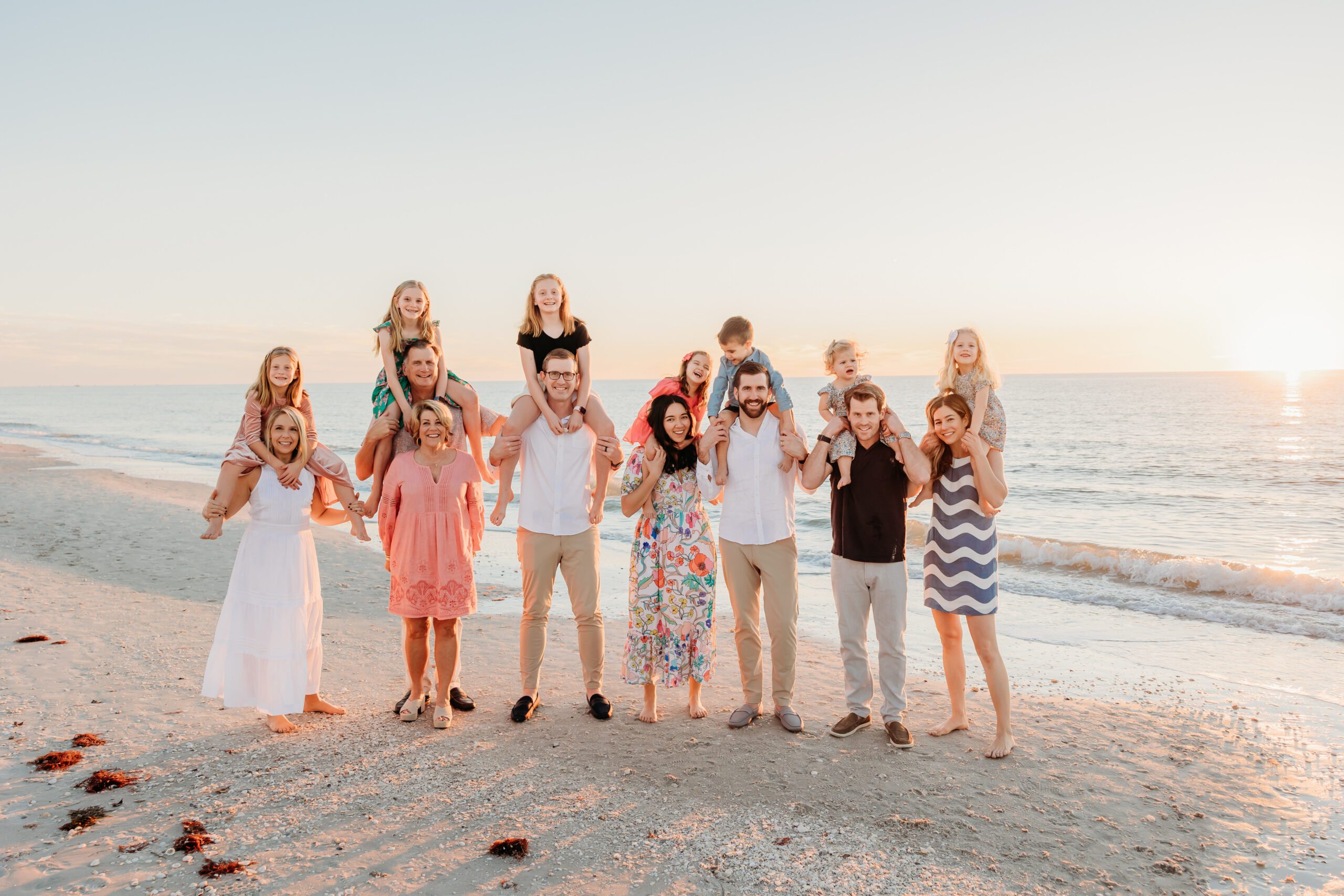 A large extended family lined up along the shoreline at sunset in Naples, parents carrying grandchildren on their shoulders as the sun drops over the Gulf